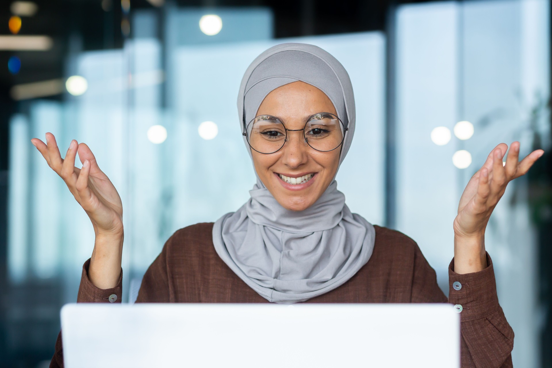 Successful and happy woman inside office at workplace, businesswoman boss in hijab talking on video call using laptop sitting at table, muslim woman joyfully gesturing to colleagues in online meeting